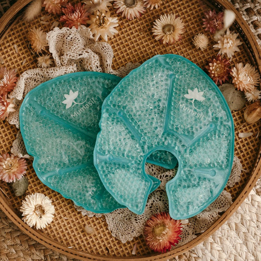 Two large, leaf-shaped, blue gel breast packs placed on a woven mat surrounded by dried flowers, presented as a product for nursing mothers.
